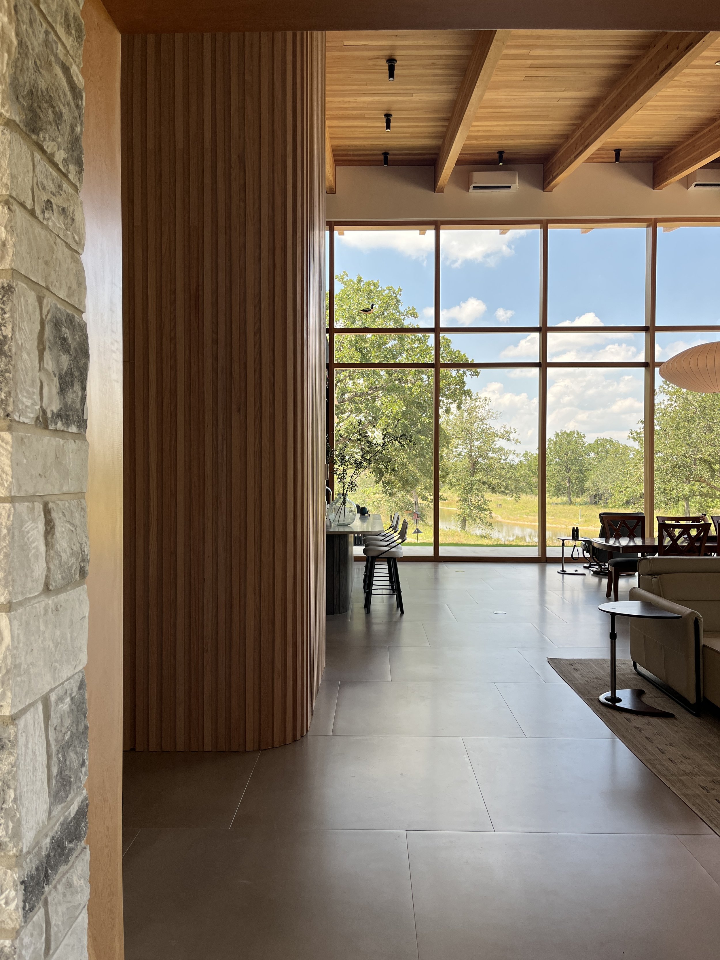 A bright, open room of a new construction home.  Large windows overlook a lake and architectural design details abound.  There is a curved wooden slat wall with hidden doors.