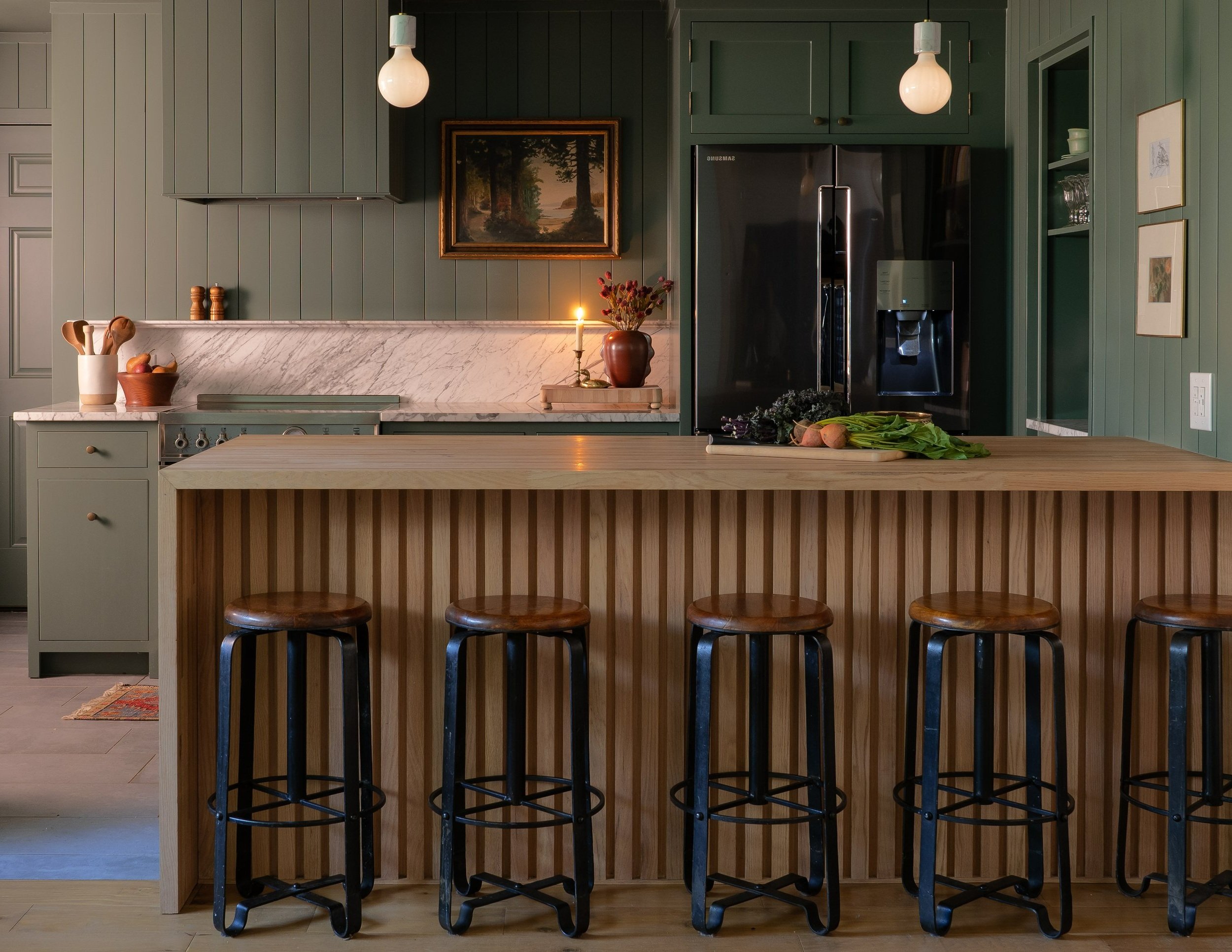 Moody green remodeled kitchen with marble and wood countertops, a lit candle, and fresh vegetables on a cutting board. A warm, calming space perfect for a luxury kitchen remodel.
